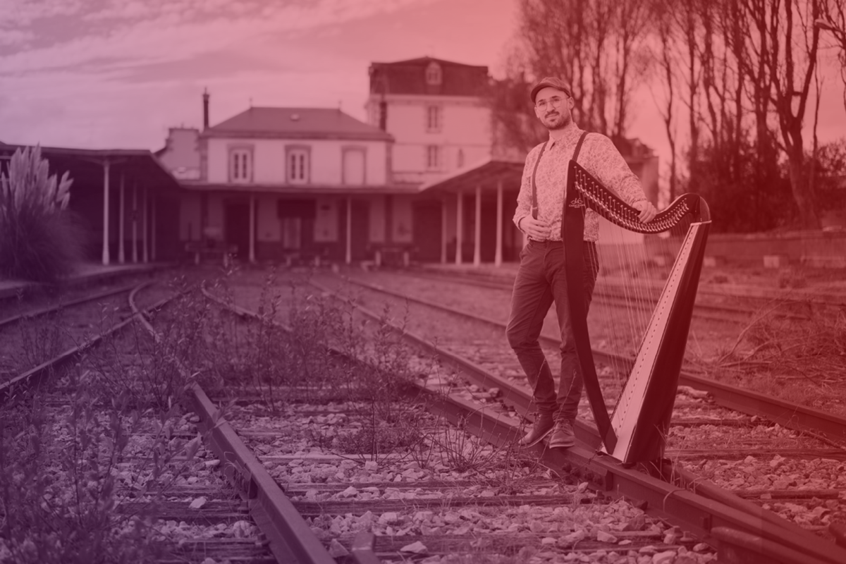 Kévin Le Pennec avec sa harpe sur une voie de chemin de fer devant une gare